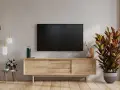 Nowoczesny salon z telewizorem na ścianie. Obok stoi drewniana szafka RTV, a na niej ozdobna roślina. W tle widać fragment ściany z fakturą przypominającą cegłę.