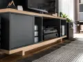 Nowoczesna szafka RTV z czarnymi frontami i drewnianymi elementami. W otwartych wnękach znajdują się sprzęty elektroniczne i płyty. Idealne miejsce, by jak schować kable.