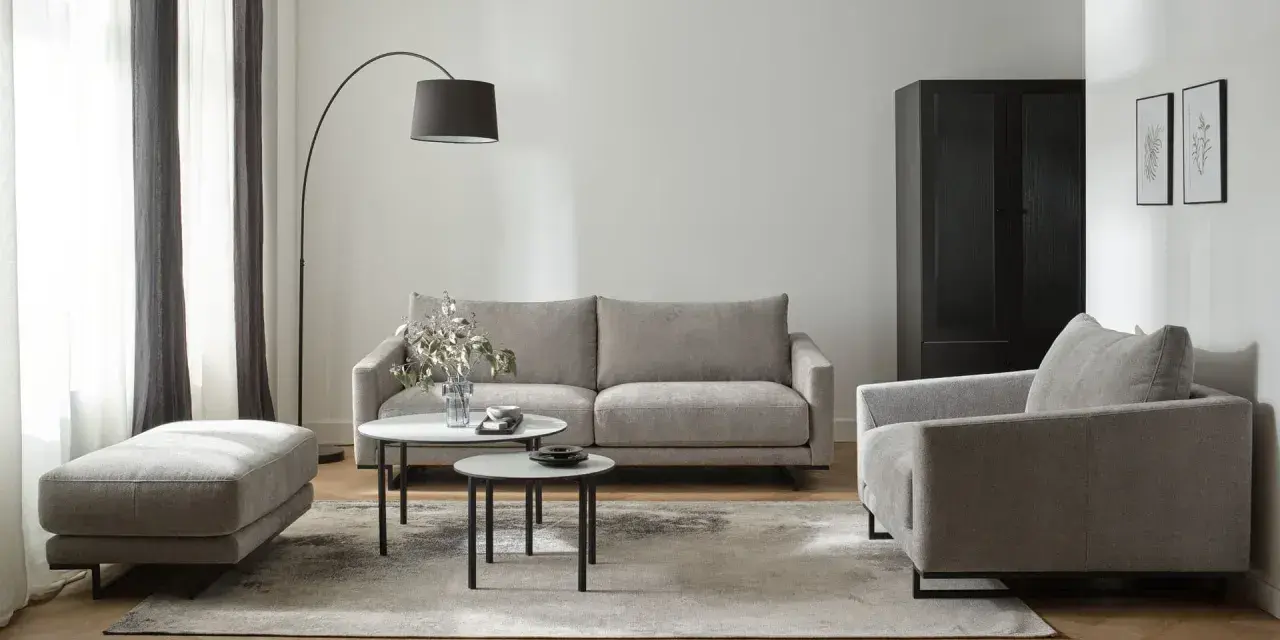 Nowoczesne biało szare salon aranżacje: szara sofa, fotel, puf i stoliki kawowe na tle białej ściany i drewnianej podłogi.