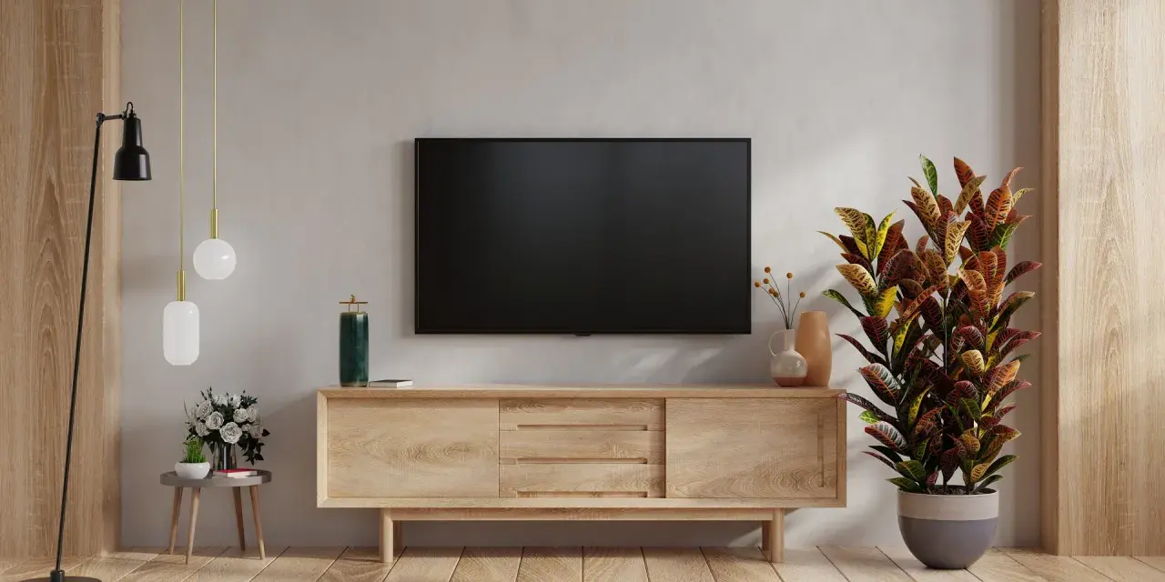 Nowoczesny salon z telewizorem na ścianie. Obok stoi drewniana szafka RTV, a na niej ozdobna roślina. W tle widać fragment ściany z fakturą przypominającą cegłę.