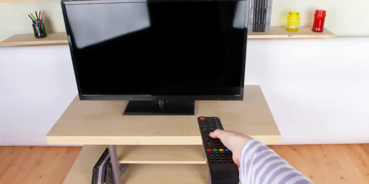 Ręka z pilotem sięga do telewizora naprzeciwko okna. Na półkach stoją płyty i kolorowe słoiki.