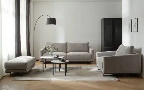 Nowoczesne biało szare salon aranżacje: szara sofa, fotel, puf i stoliki kawowe na tle białej ściany i drewnianej podłogi.
