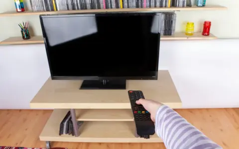 Ręka z pilotem sięga do telewizora naprzeciwko okna. Na półkach stoją płyty i kolorowe słoiki.