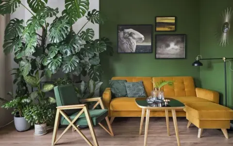 Zielony kącik z kwiatami w salonie. Monstera i inne rośliny tworzą bujną dżunglę obok musztardowej kanapy i fotela.