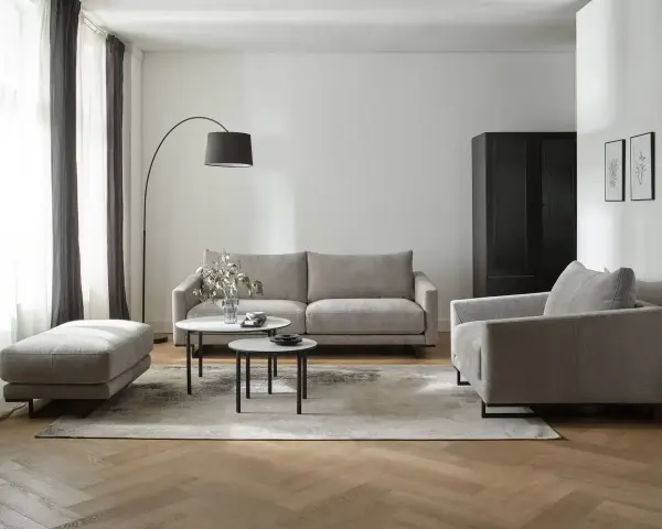 Nowoczesne biało szare salon aranżacje: szara sofa, fotel, puf i stoliki kawowe na tle białej ściany i drewnianej podłogi.