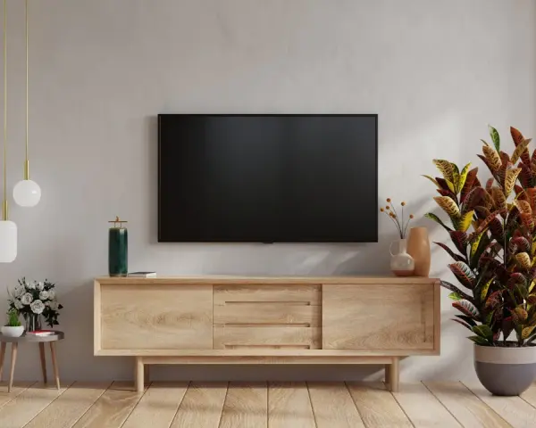Nowoczesny salon z telewizorem na ścianie. Obok stoi drewniana szafka RTV, a na niej ozdobna roślina. W tle widać fragment ściany z fakturą przypominającą cegłę.