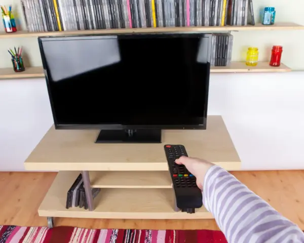 Ręka z pilotem sięga do telewizora naprzeciwko okna. Na półkach stoją płyty i kolorowe słoiki.