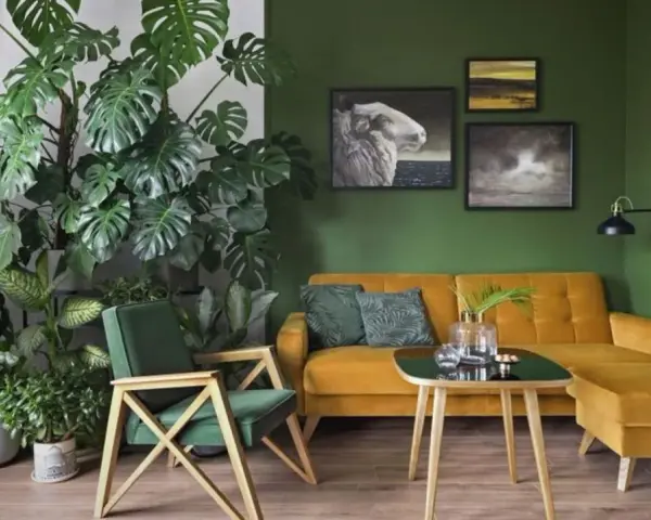 Zielony kącik z kwiatami w salonie. Monstera i inne rośliny tworzą bujną dżunglę obok musztardowej kanapy i fotela.