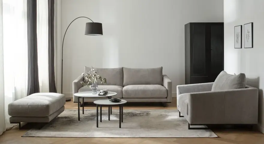 Nowoczesne biało szare salon aranżacje: szara sofa, fotel, puf i stoliki kawowe na tle białej ściany i drewnianej podłogi.