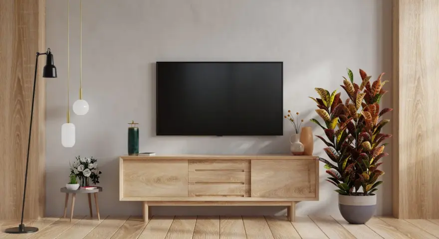 Nowoczesny salon z telewizorem na ścianie. Obok stoi drewniana szafka RTV, a na niej ozdobna roślina. W tle widać fragment ściany z fakturą przypominającą cegłę.
