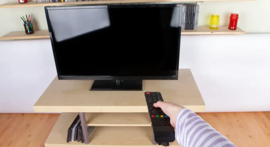 Ręka z pilotem sięga do telewizora naprzeciwko okna. Na półkach stoją płyty i kolorowe słoiki.