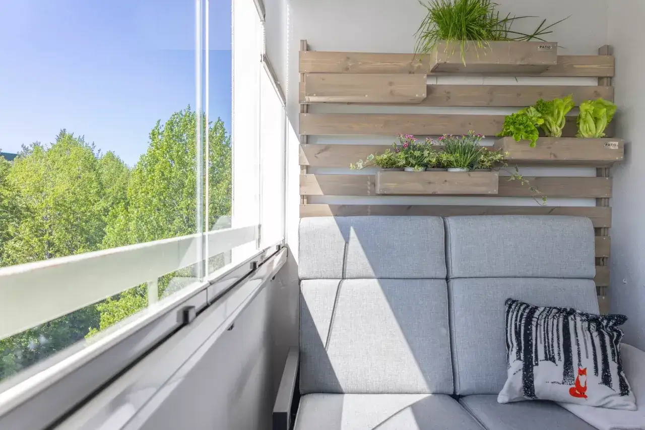 Przytulny balkon tanim kosztem z szarą sofą, poduszką z liskiem i zielenią na drewnianej ściance.