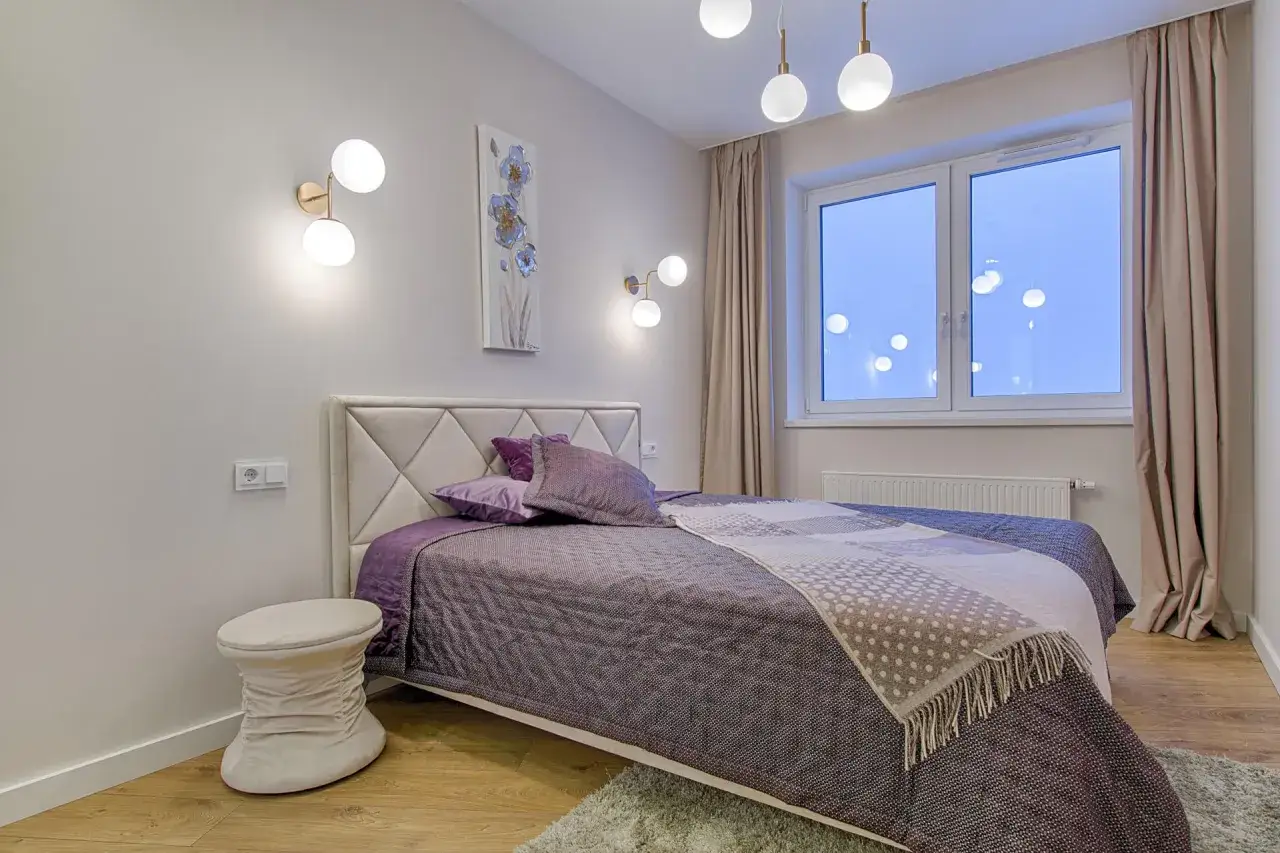 Elegancka sypialnia z łóżkiem, na którym znajduje się narzuta w rozmiarze 160x200. Wygodny puf i stylowe oświetlenie dopełniają aranżacji.