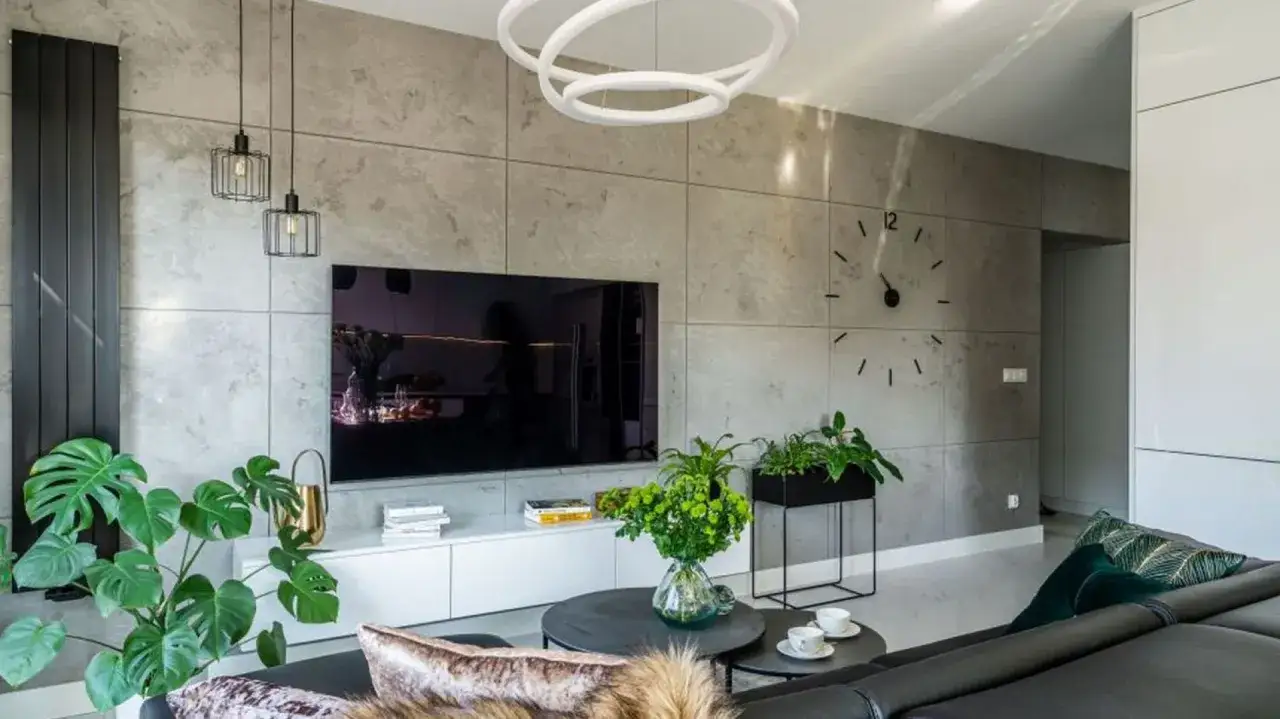 Nowoczesny salon z dużym telewizorem naprzeciwko okna. Ściana za TV ozdobiona betonem architektonicznym, a obok zegar.
