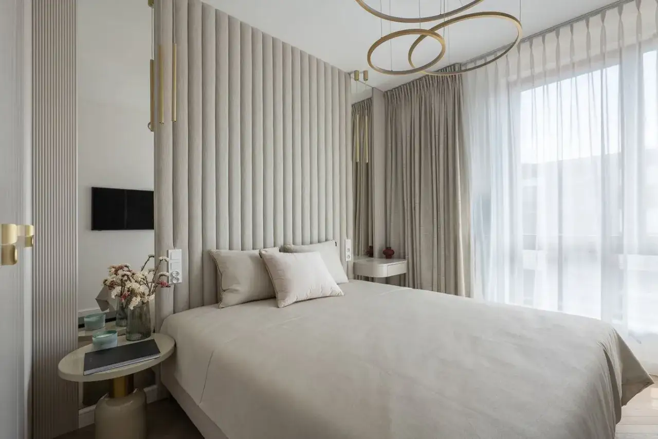 Elegancka aranzacja sypialni z tapicerowanym zagłówkiem, lustrem i nowoczesnym oświetleniem. Miękkie tkaniny i stonowana kolorystyka tworzą przytulną atmosferę.